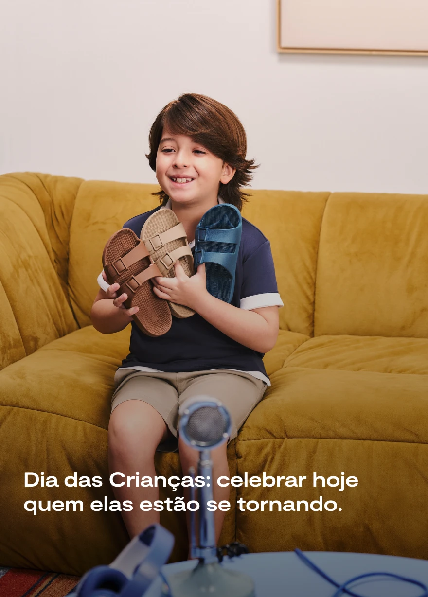 Menino sentado em sofá amarelo segurando três sandálias Linus coloridas Aramis Next e Linus.